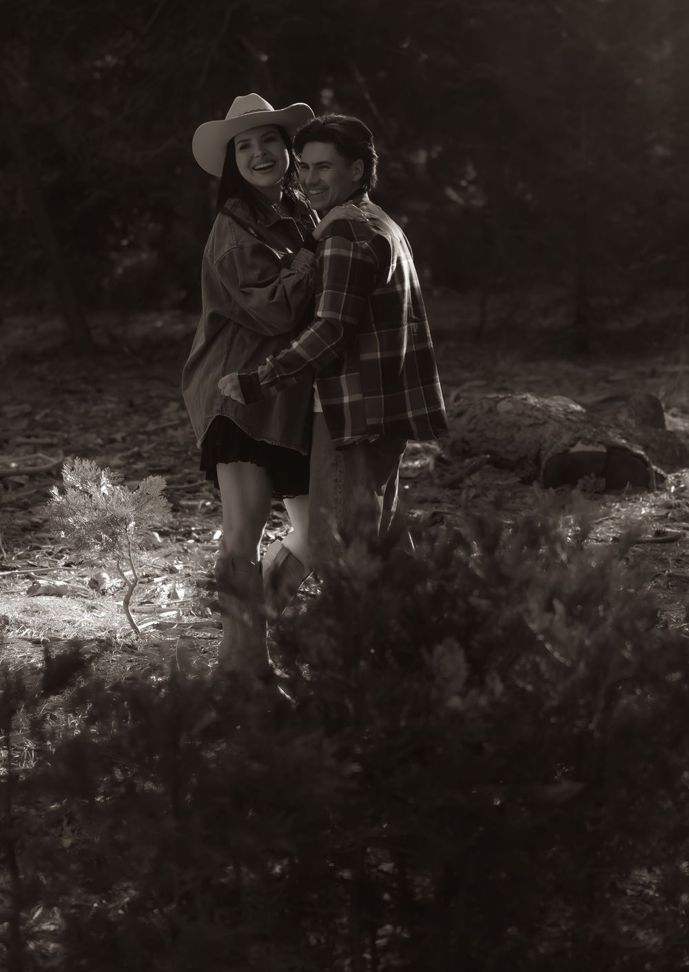 Two figures sit together in a romantic black and white scene on a fallen tree trunk in a forest setting.