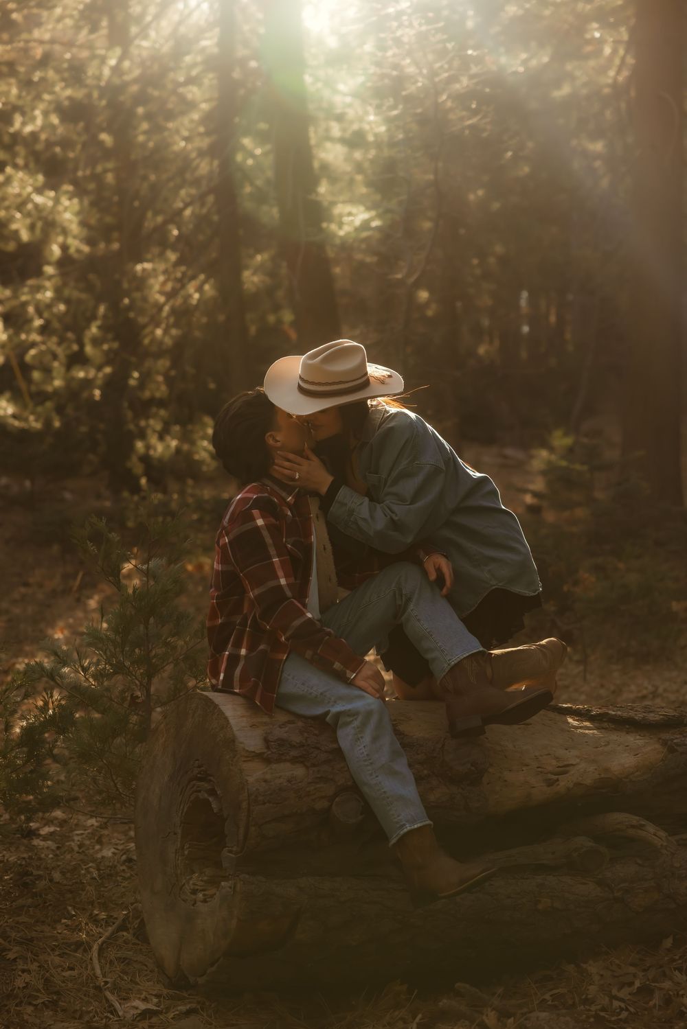 A person in cowboy attire sits on the ground playing an acoustic guitar in a sunlit forest setting.