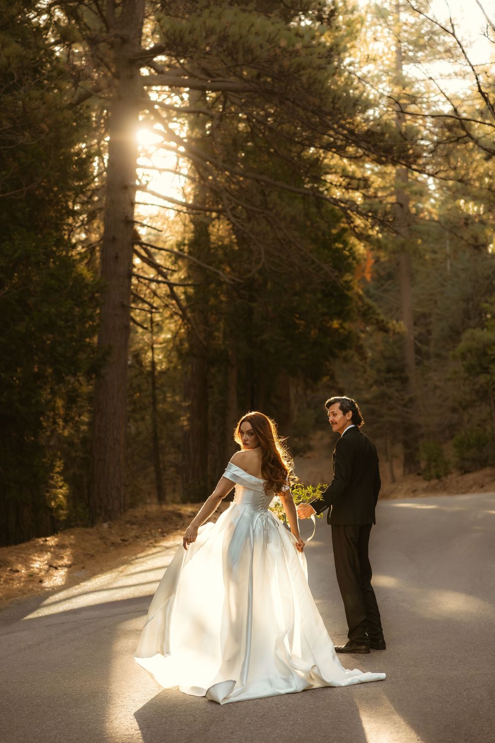 A couple in wedding attire dances together on a snowy path at sunset with sunlight filtering through pine trees.