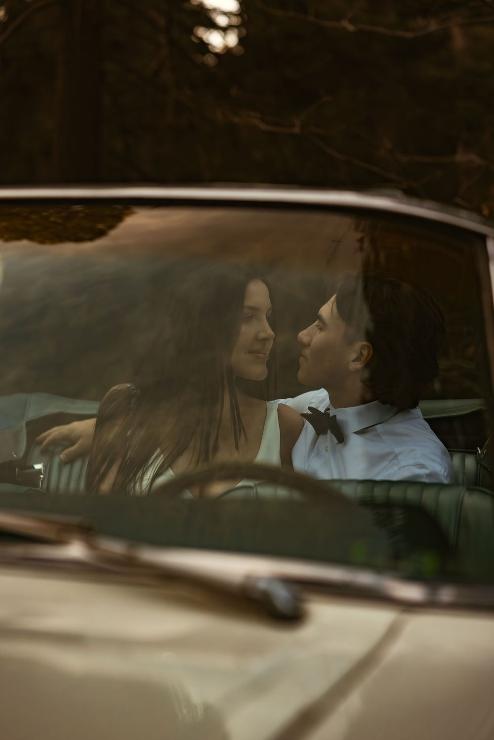 Two people sitting in a car having a conversation viewed through the windshield at dusk.