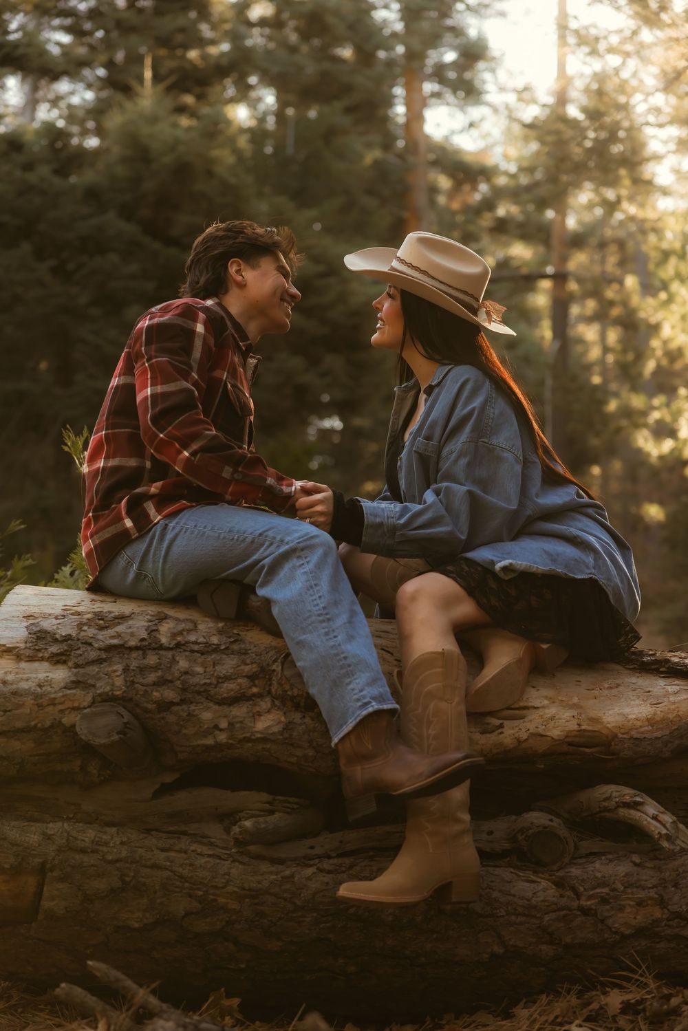 A couple in casual western attire sits together on a fallen log in the forest during golden hour.