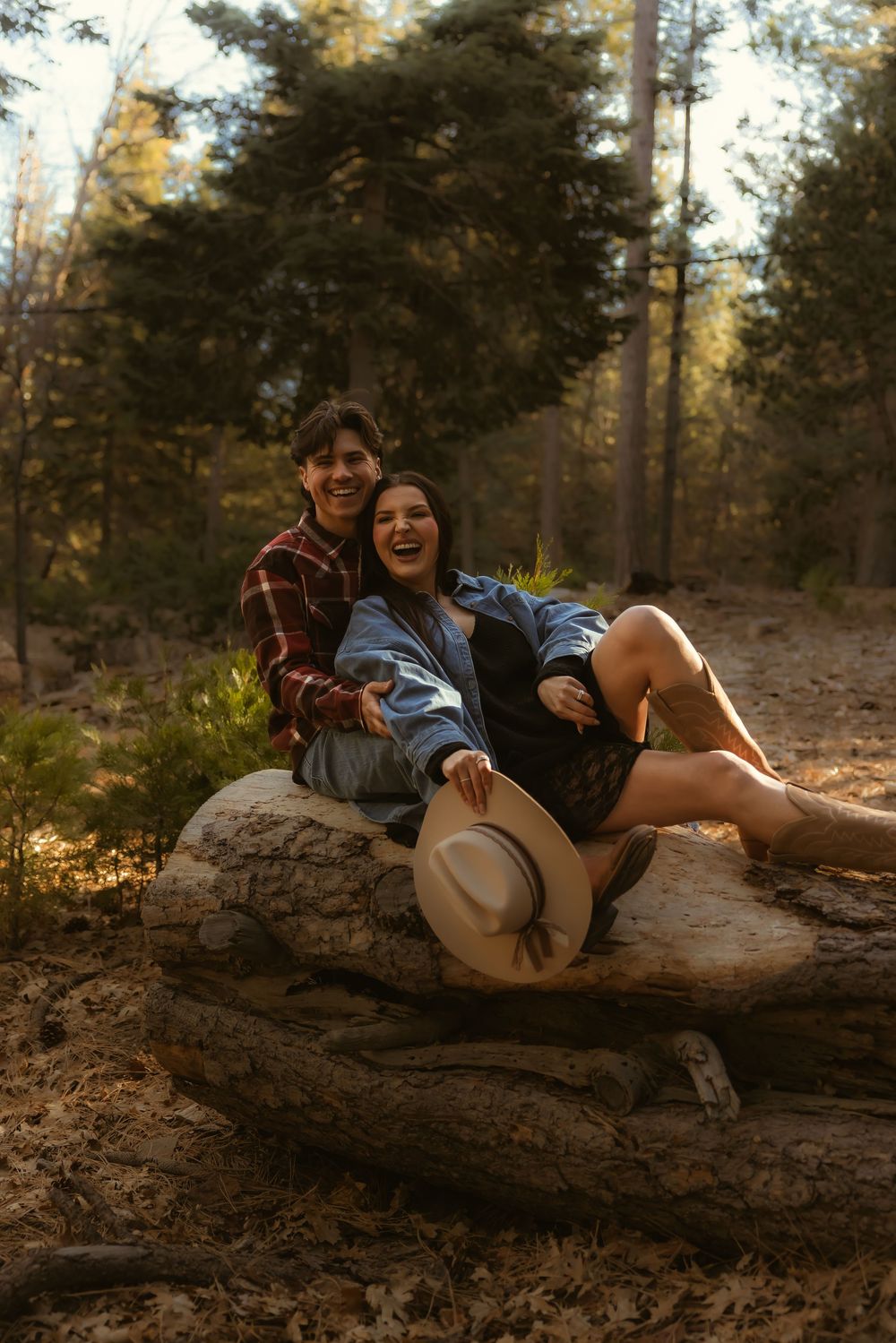 A couple sits together on a fallen log in a forest during golden hour, with a hat resting beside them.