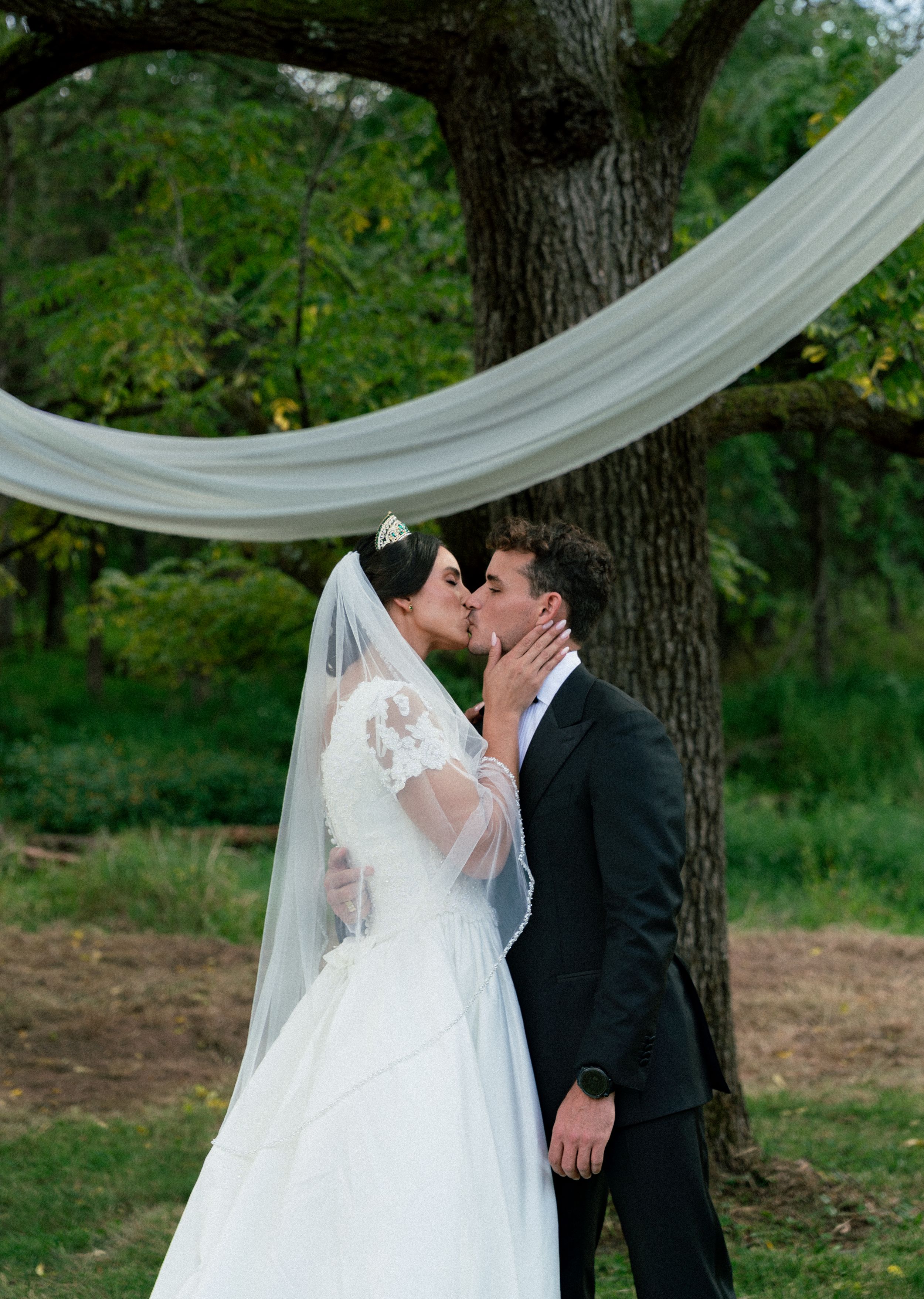 A romantic wedding moment captured under trees with flowing white veil and formal attire.