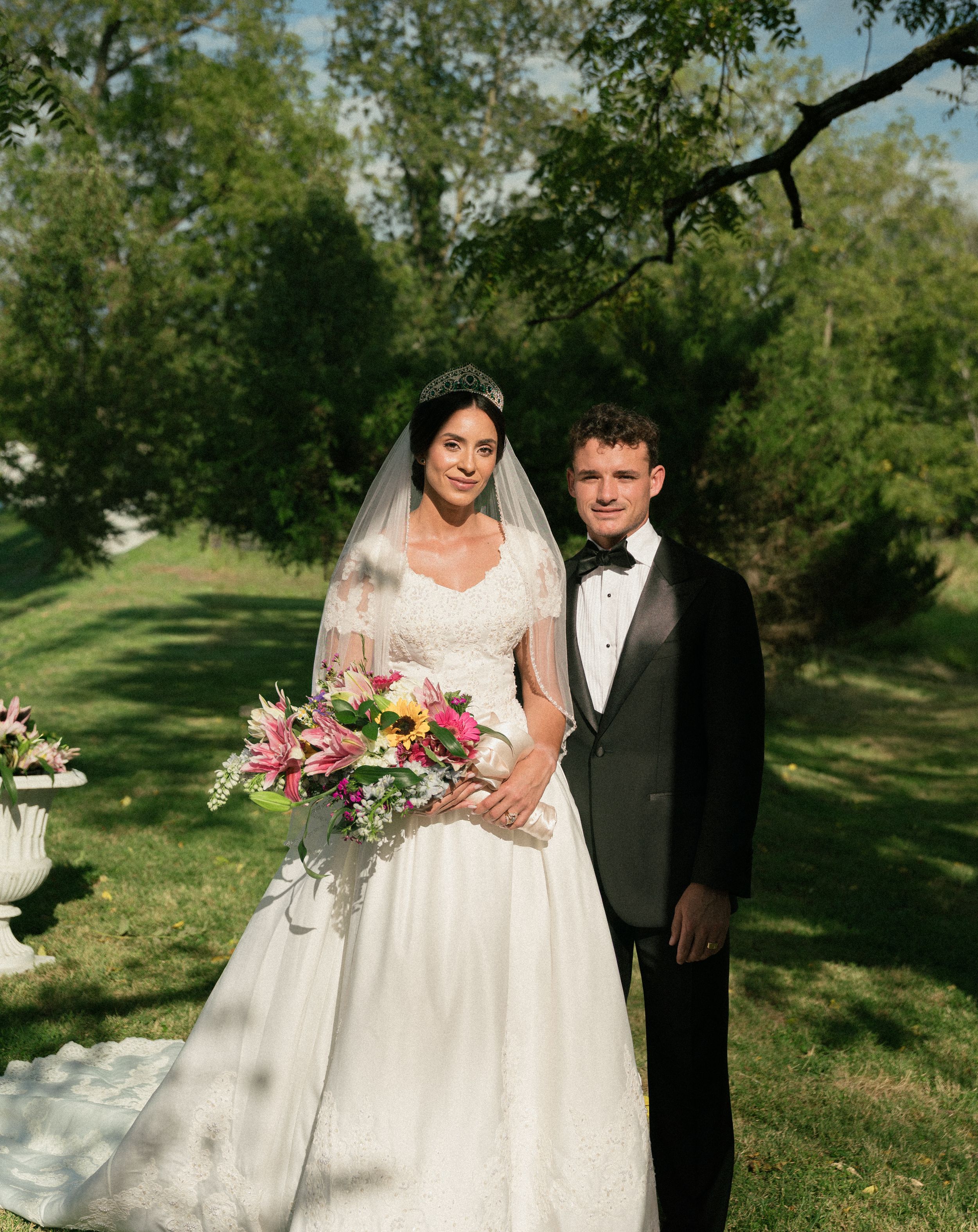 A wedding couple poses with pink bouquet in a sunny garden setting.
