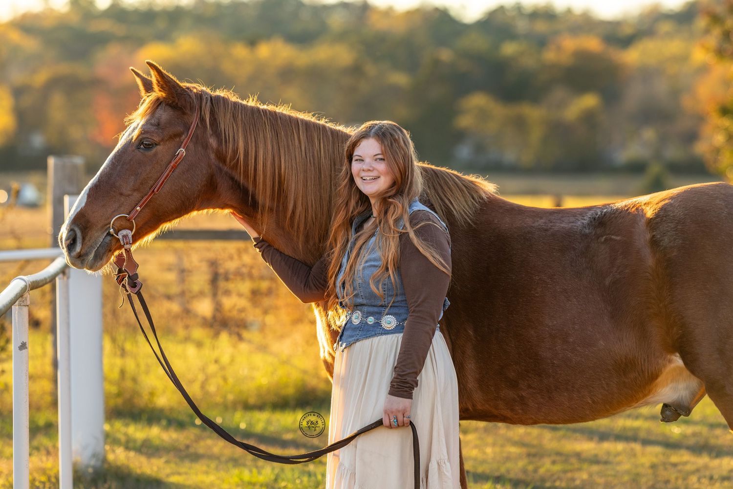 A young equestrian stands with a brown horse in a golden sunlit field with white fencing during sunset.