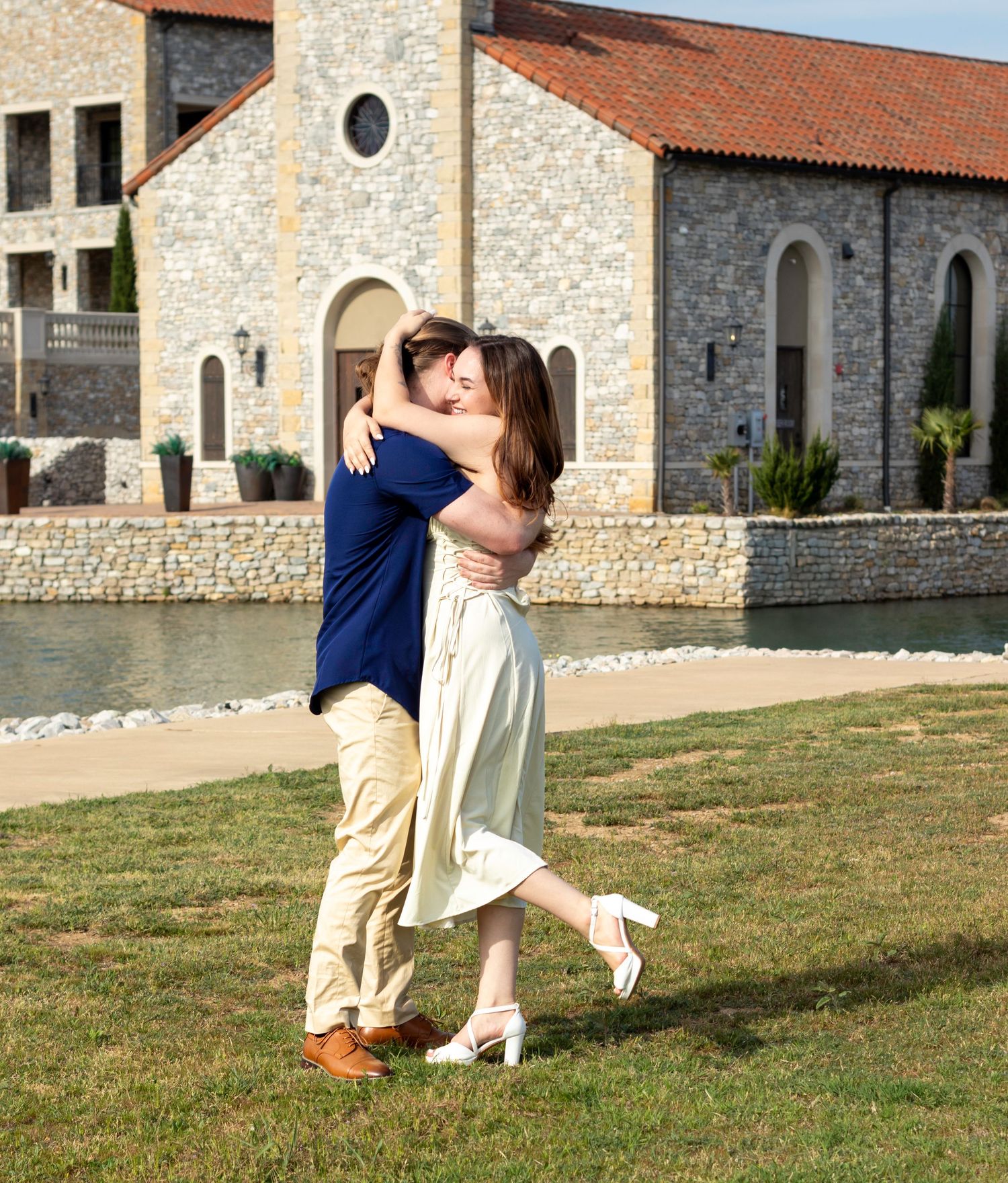 A romantic couple shares a tender dip pose on a grassy lawn in front of a rustic stone building with terracotta roof.