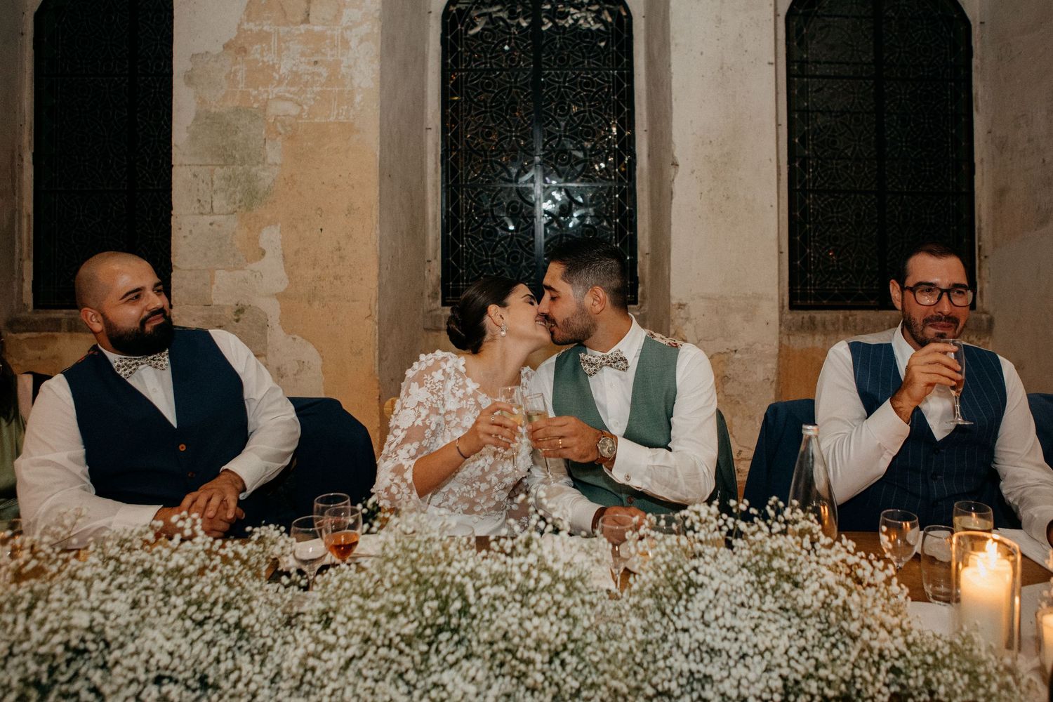 Bride and groom share a kiss at candlelit head table adorned with baby's breath flowers during wedding reception.