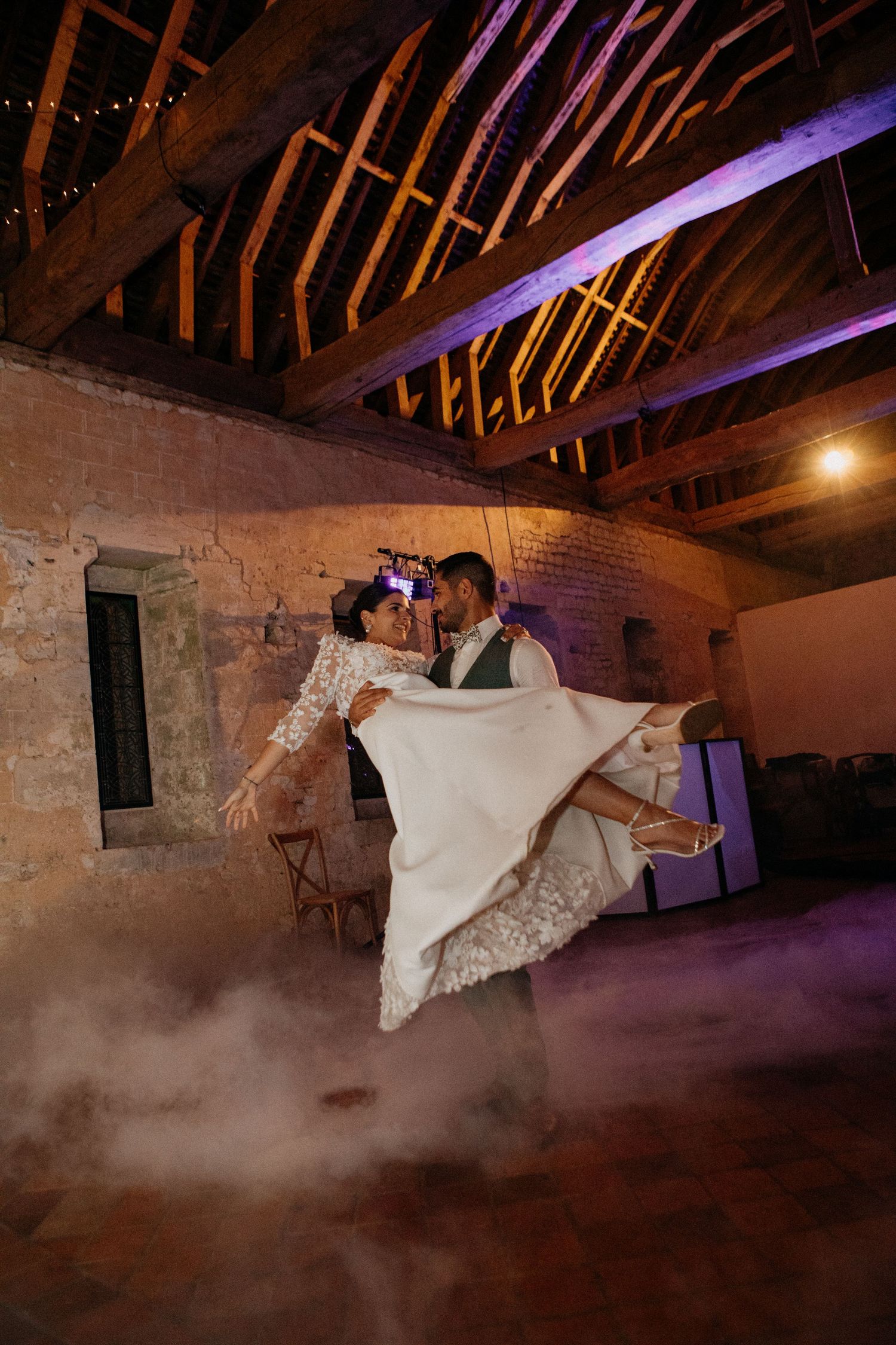 Bride and groom share romantic first dance in rustic barn venue with dramatic fog and warm lighting.
