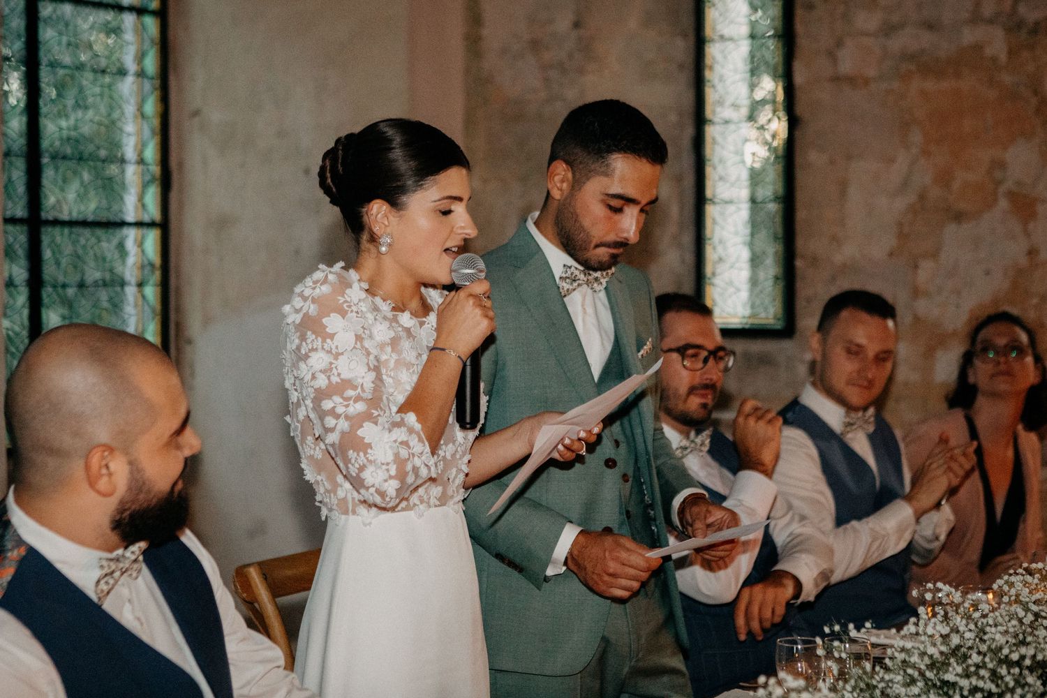 Bride in lace gown reads vows into microphone beside groom in sage green suit during intimate wedding ceremony.