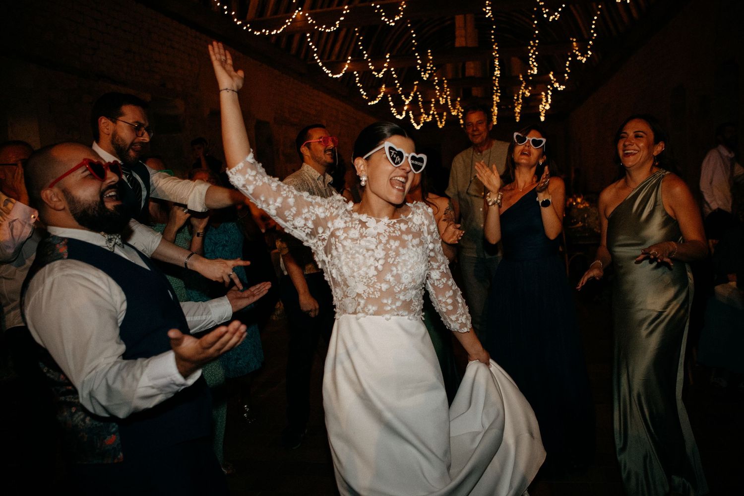 Bride in lace gown dances joyfully wearing heart-shaped sunglasses at lively wedding reception under fairy lights.