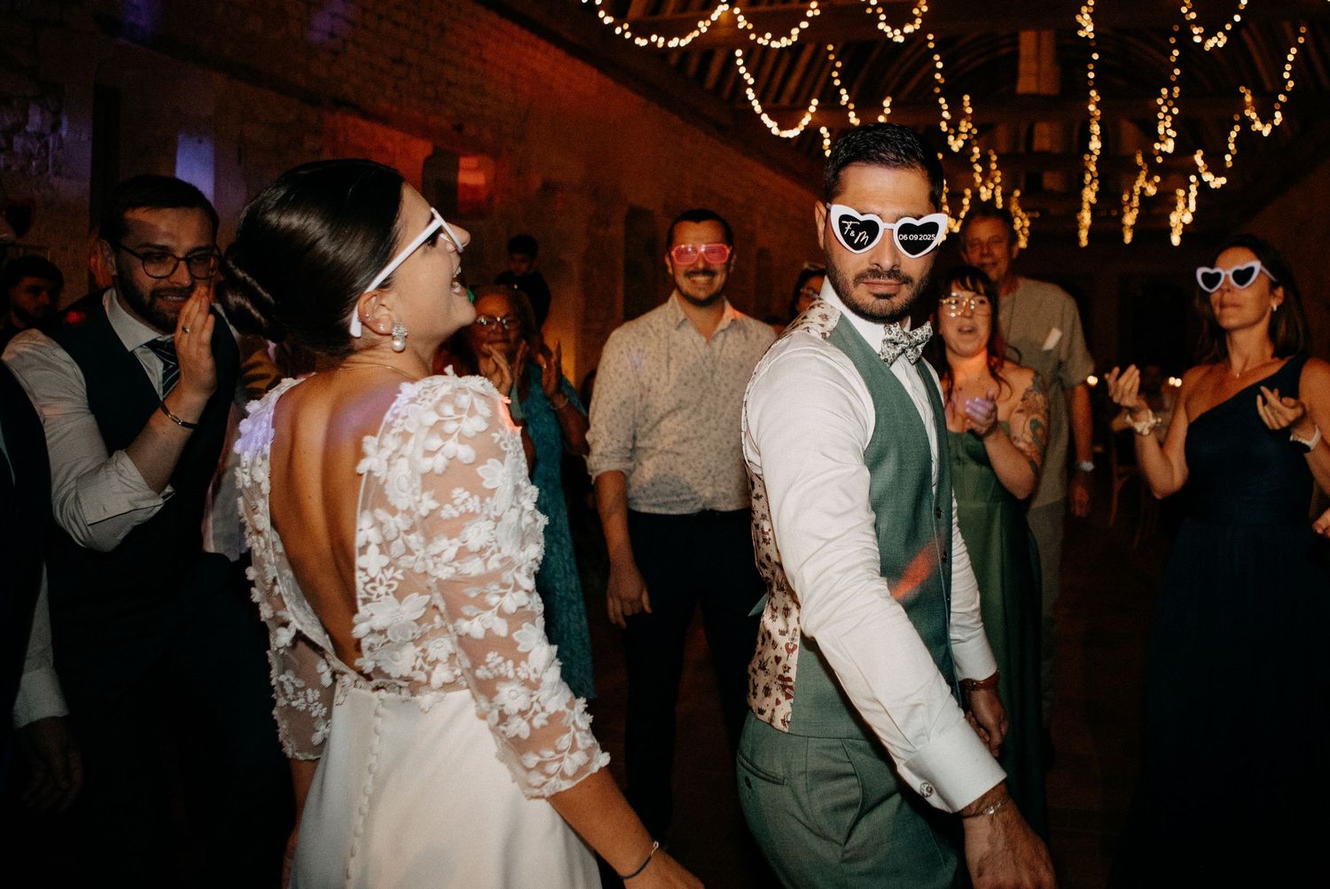 Bride and groom dance together wearing novelty heart-shaped sunglasses surrounded by guests at wedding reception.