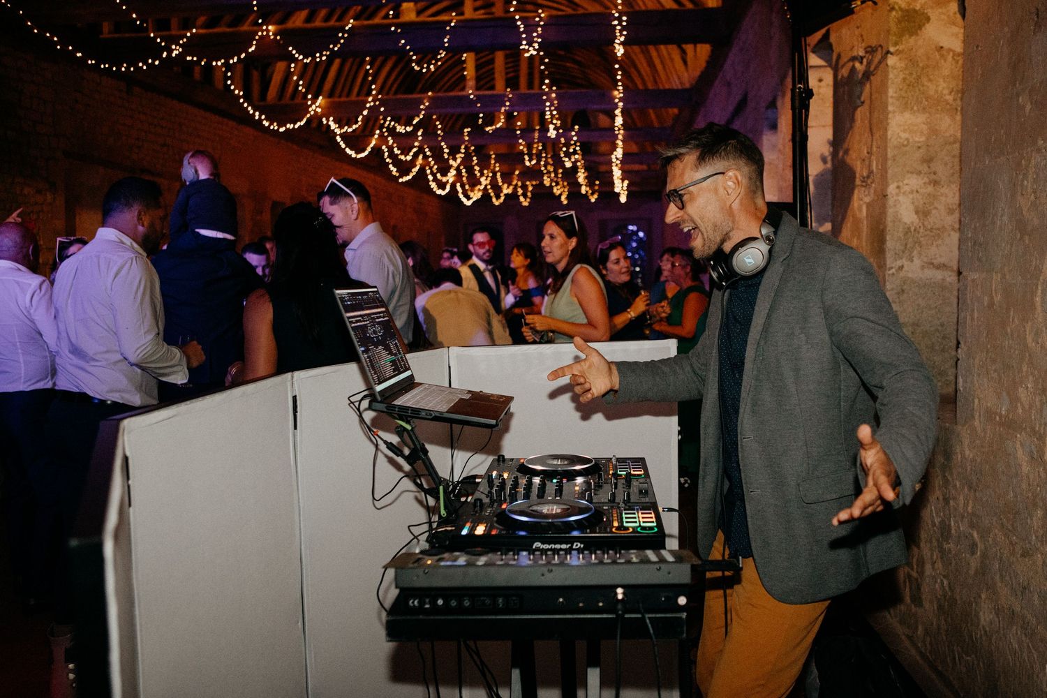 DJ performing at lively wedding reception under fairy lights in a rustic stone barn venue.