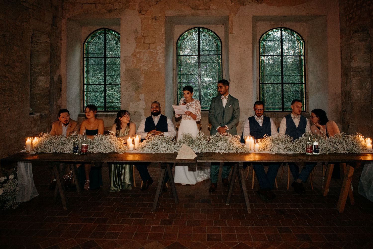 Wedding party seated at long candlelit table decorated with baby's breath in an elegant rustic stone venue.