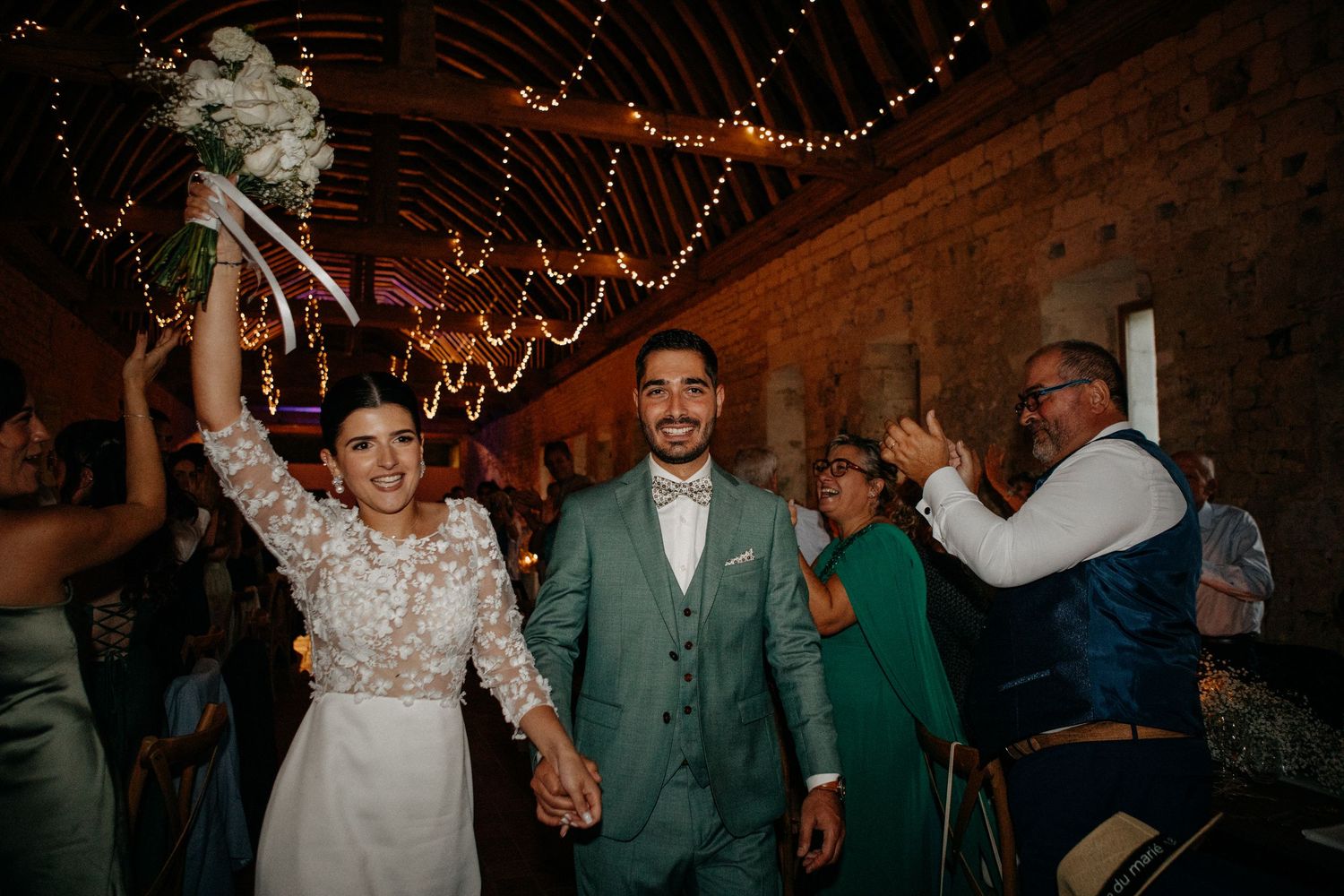 Joyful newlyweds walk hand in hand as bride raises bouquet triumphantly under fairy lights in rustic barn reception.