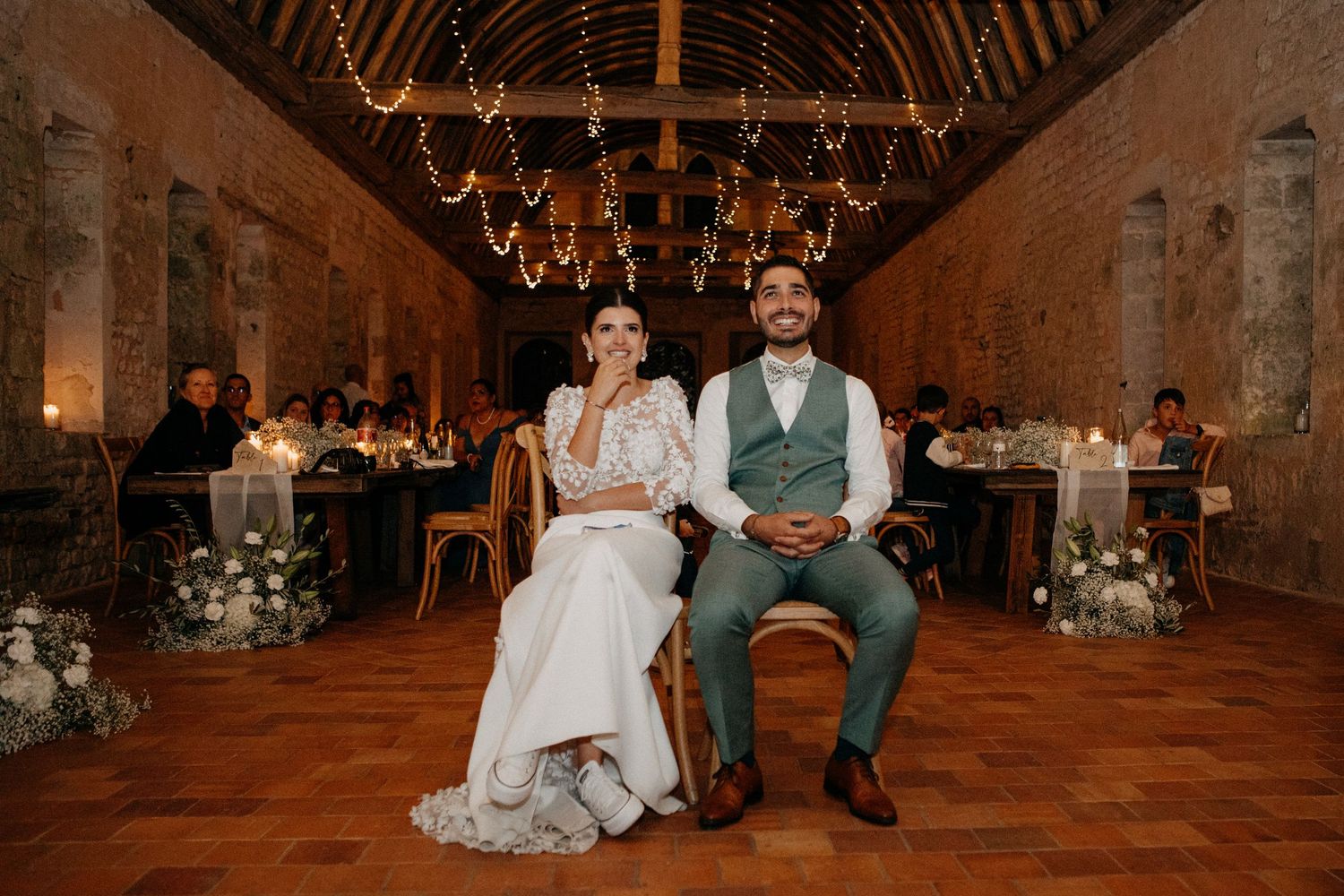 Bride and groom sit together smiling in rustic barn reception hall decorated with fairy lights and candles.