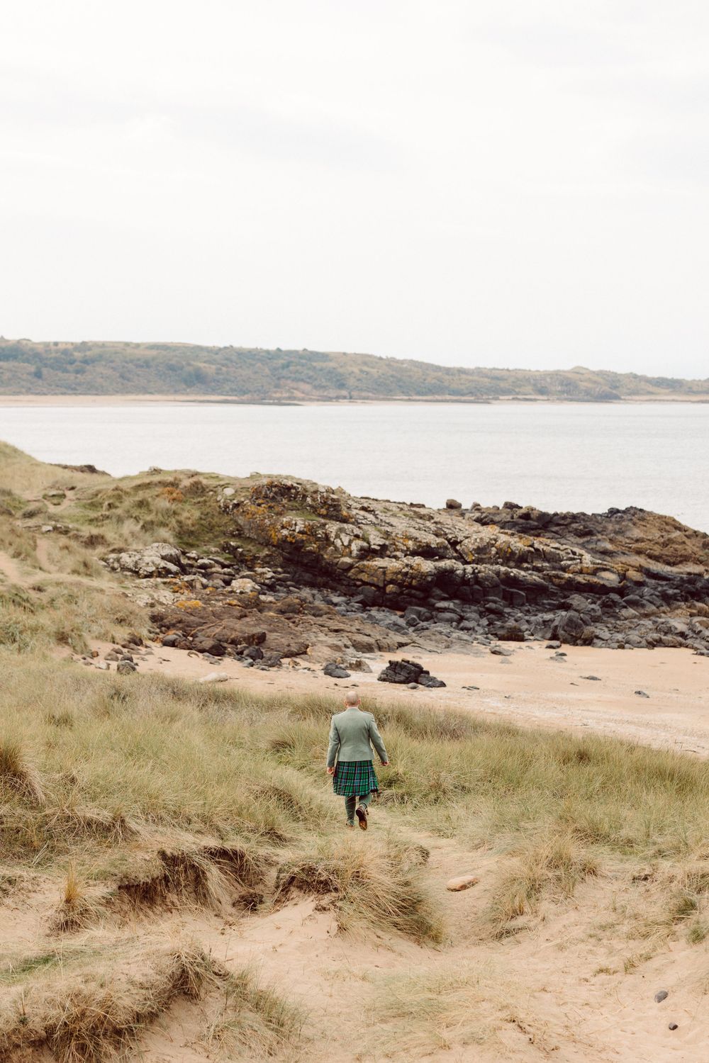 A scenic coastal path winds through grassy dunes and rocky shoreline with distant cliffs on the horizon.
