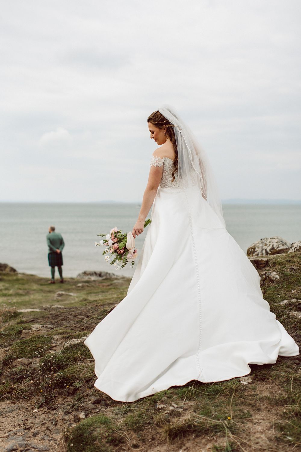 A bride in a flowing white wedding dress walks along a coastal cliff edge with dramatic ocean views in the background.