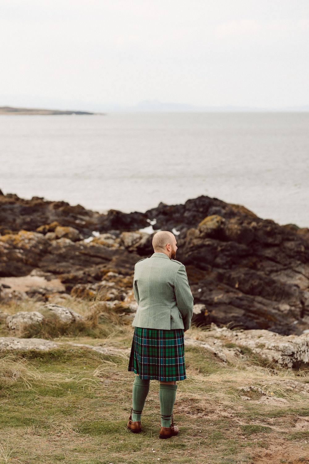Person in traditional Scottish kilt and green jacket sits contemplatively on coastal cliffs overlooking the misty ocean.