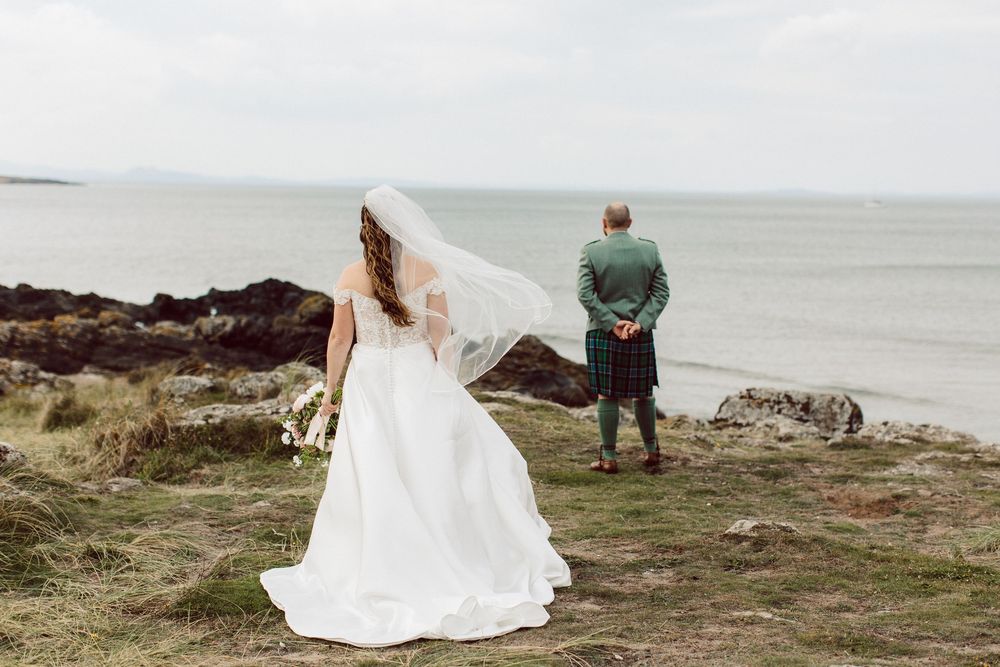 Couple in wedding attire sits on coastal rocks overlooking a misty ocean view.
