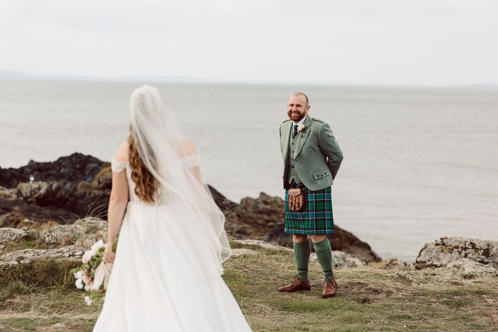 A wedding couple walks along a misty coastal cliff with dramatic ocean views and rocky outcrops in the background.