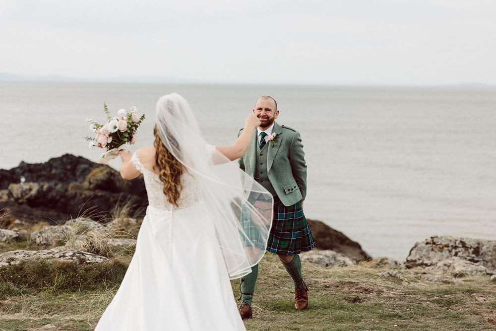A romantic coastal wedding moment captured on a rocky beach with waves and cliffs in the background.