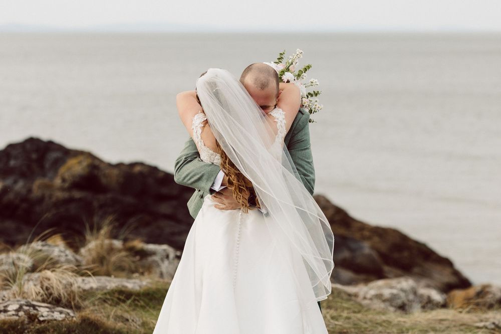 A bride in a flowing white wedding dress stands gracefully on a rocky coastal shoreline during sunset.