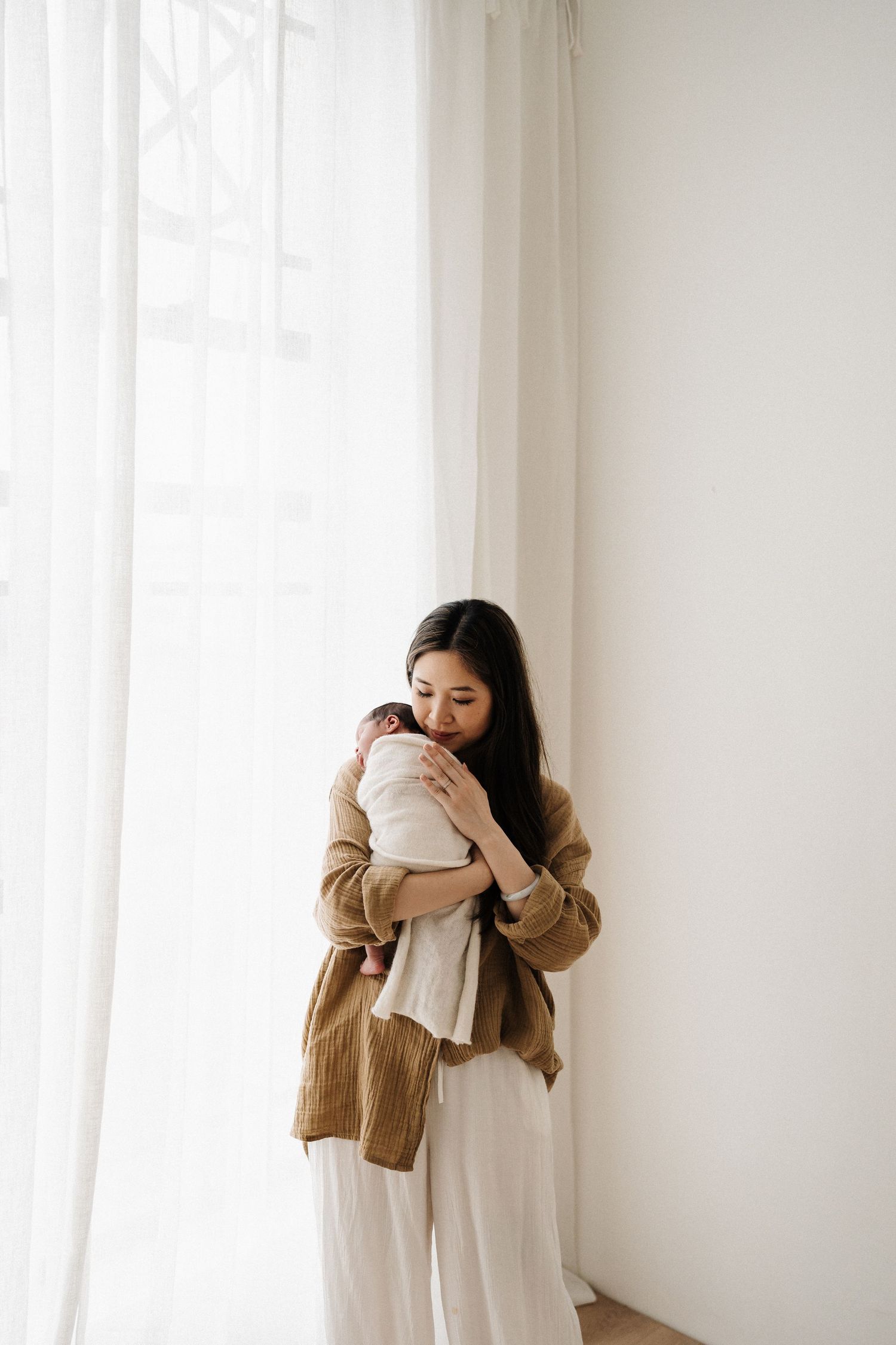 A person in a cozy brown cardigan and white pants holds a baby near a bright window with sheer curtains.