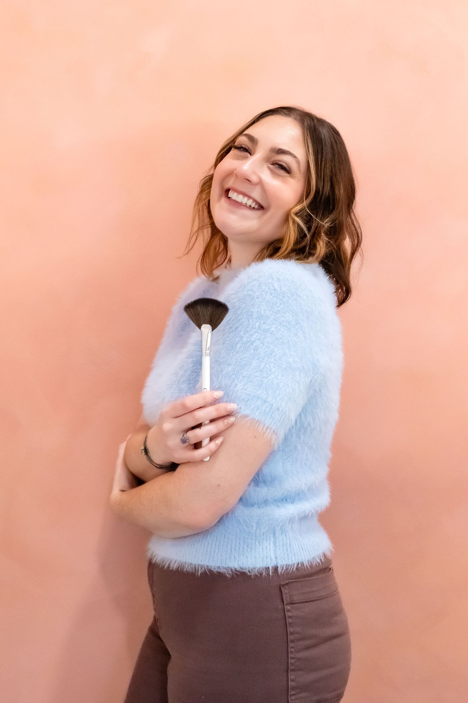 A smiling person in a fuzzy blue sweater holds a makeup brush while posing against a peach-colored wall.