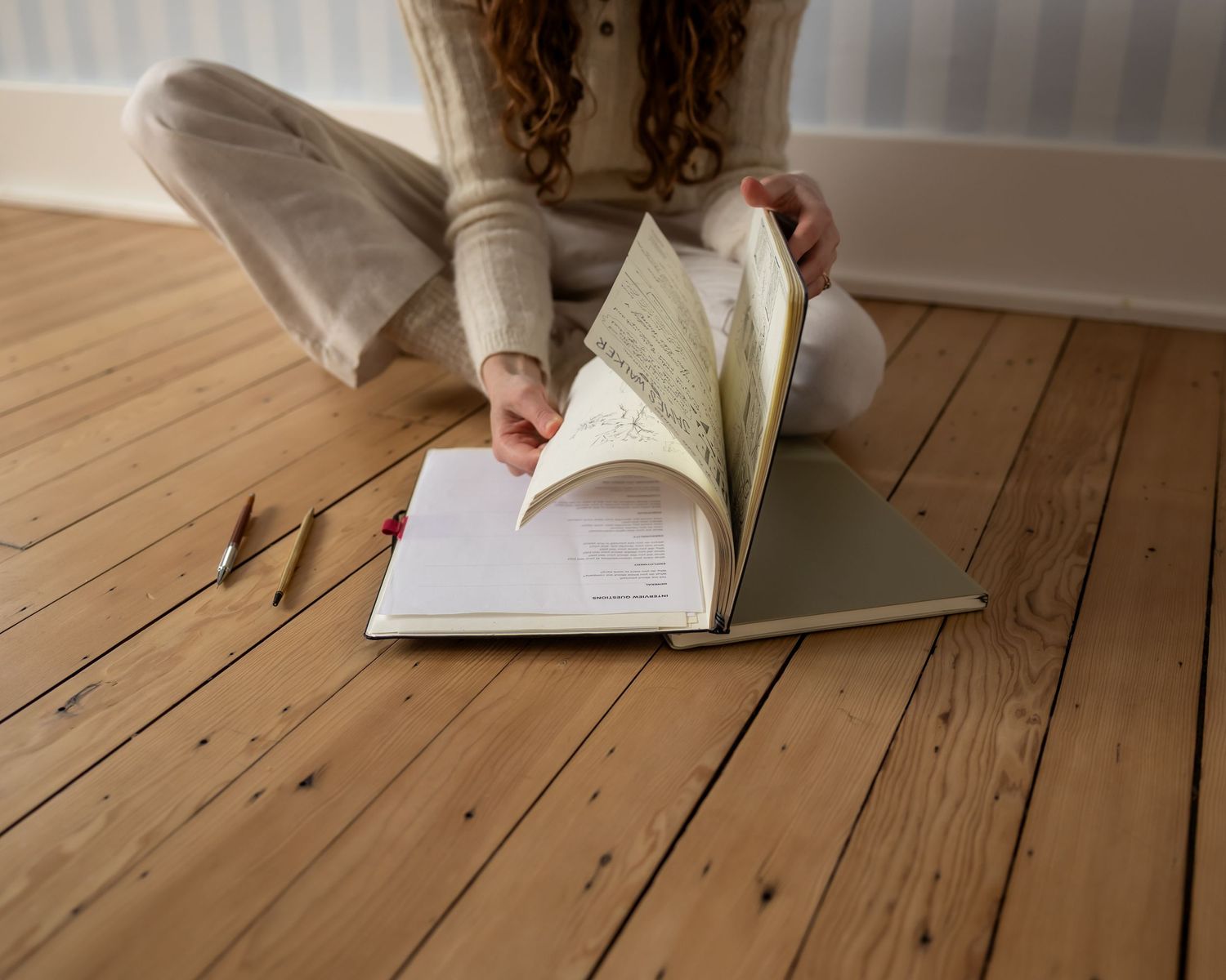 A hand holding a pen writes in a notebook on a rustic wooden floor with natural lighting.