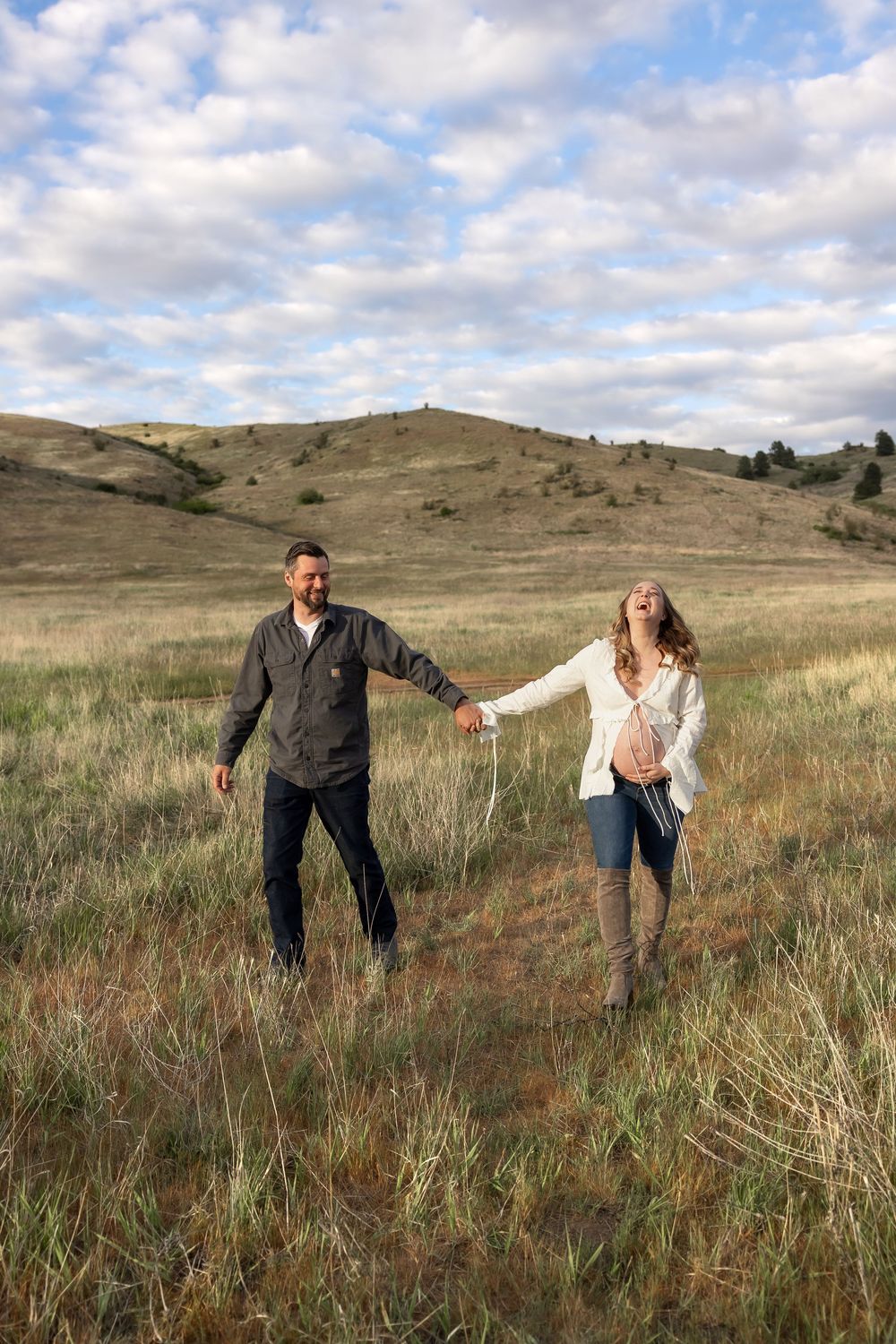 A couple holds hands while standing apart in a grassy hillside meadow under cloudy skies.