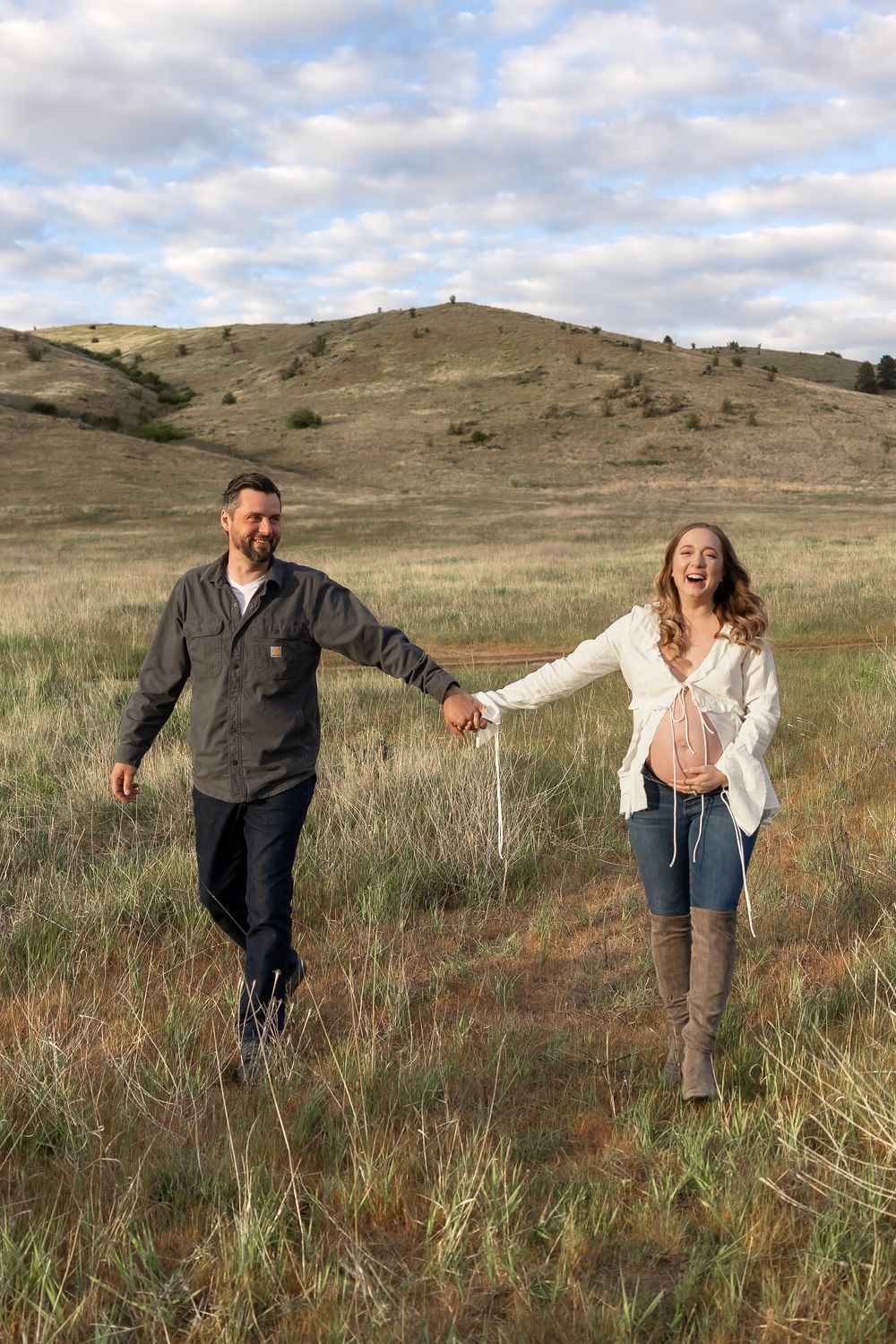 A couple walks hand in hand through a grassy hillside field on a cloudy day.