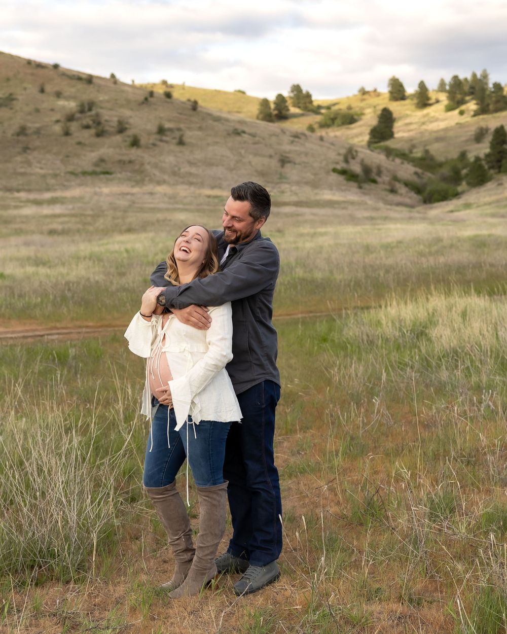 A couple embraces while laughing together in a scenic grassy hillside landscape during golden hour.