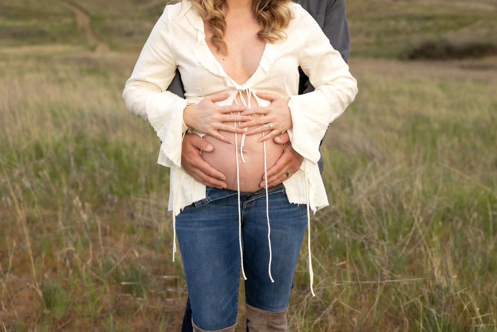 A couple stands together in a grassy field wearing casual attire on a sunny day.