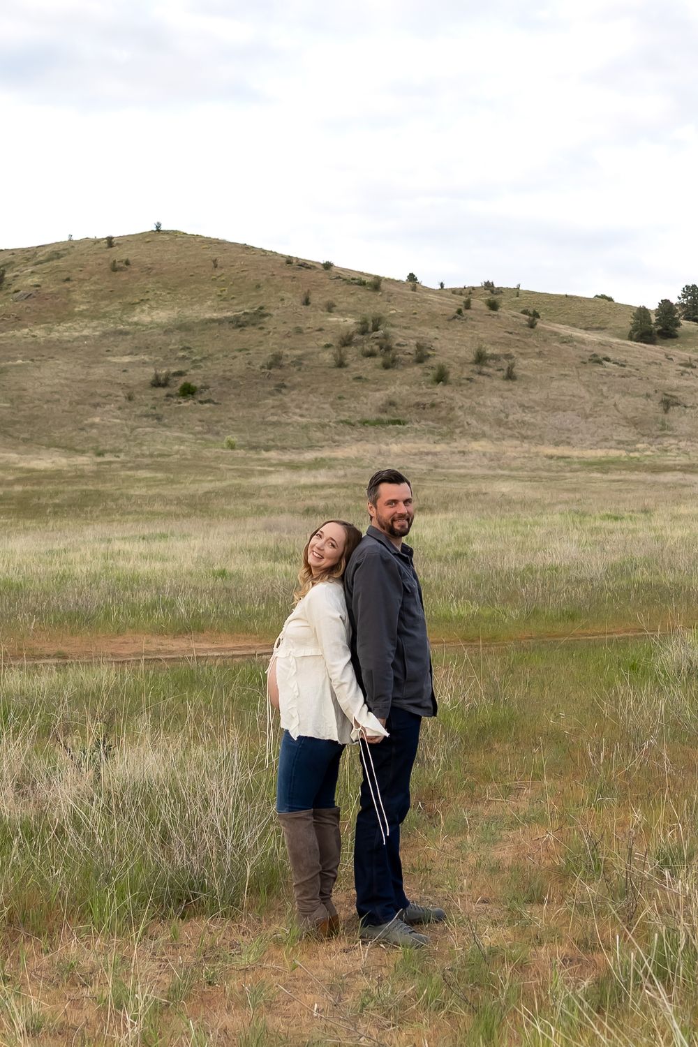 A couple standing back-to-back in a grassy meadow with a scenic hillside behind them.