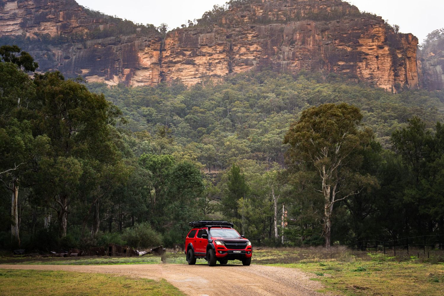 Red 4WD vehicle driving on dirt road through Australian bushland with dramatic sandstone cliff faces in background.