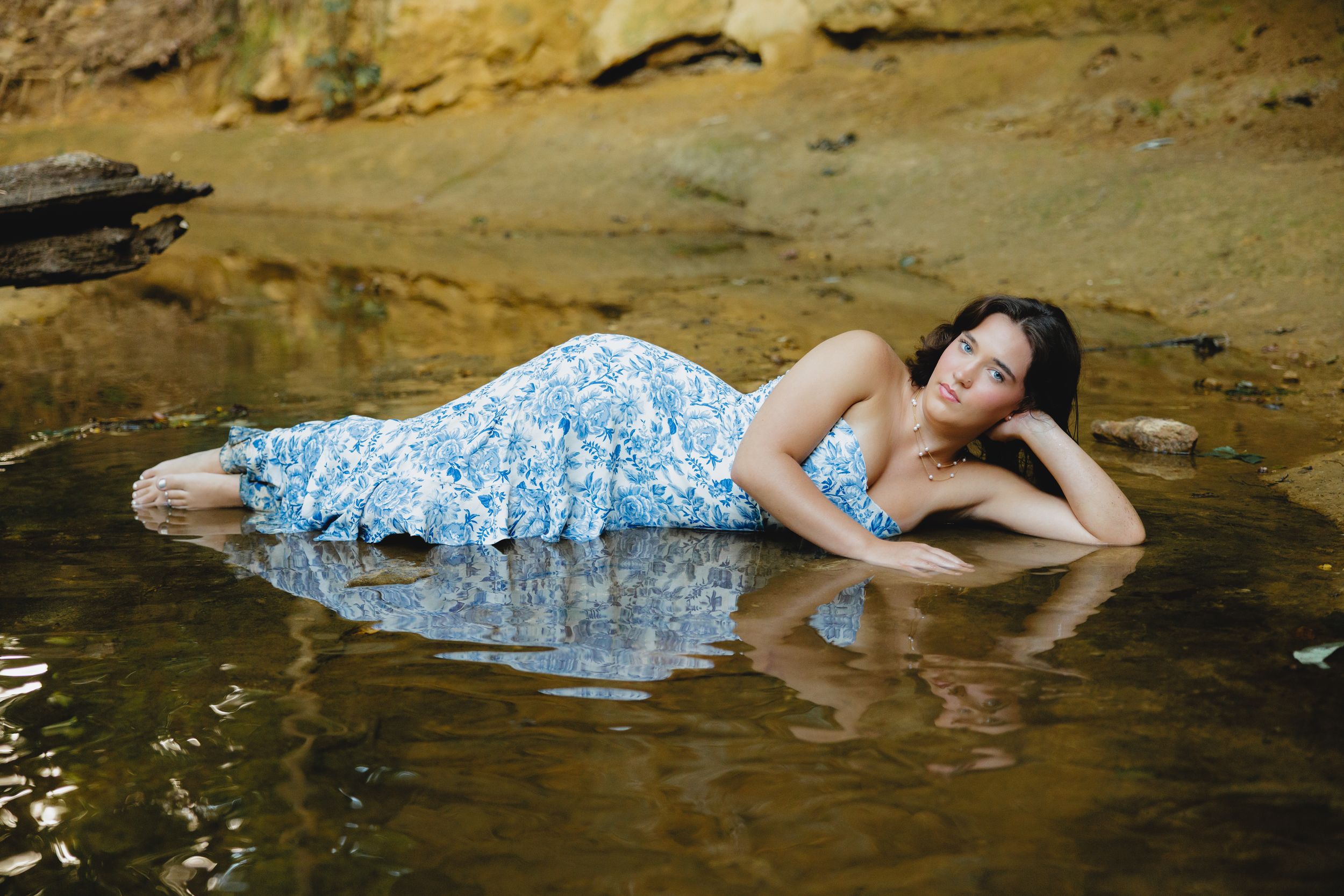 A senior in a blue floral dress photographed in shallow water with artistic reflection.
