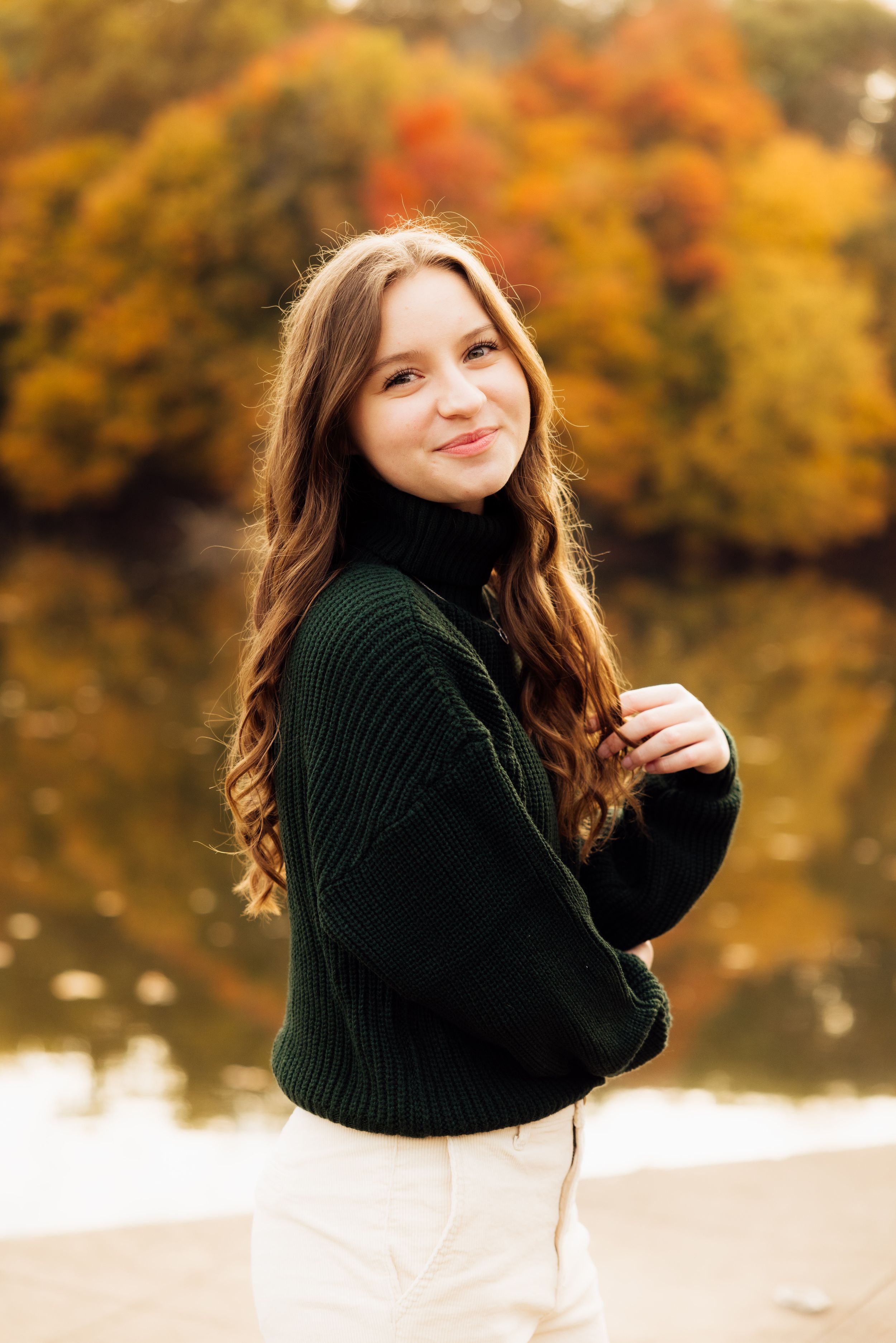 Fall senior portrait session by a lake with warm autumn colors in the background.