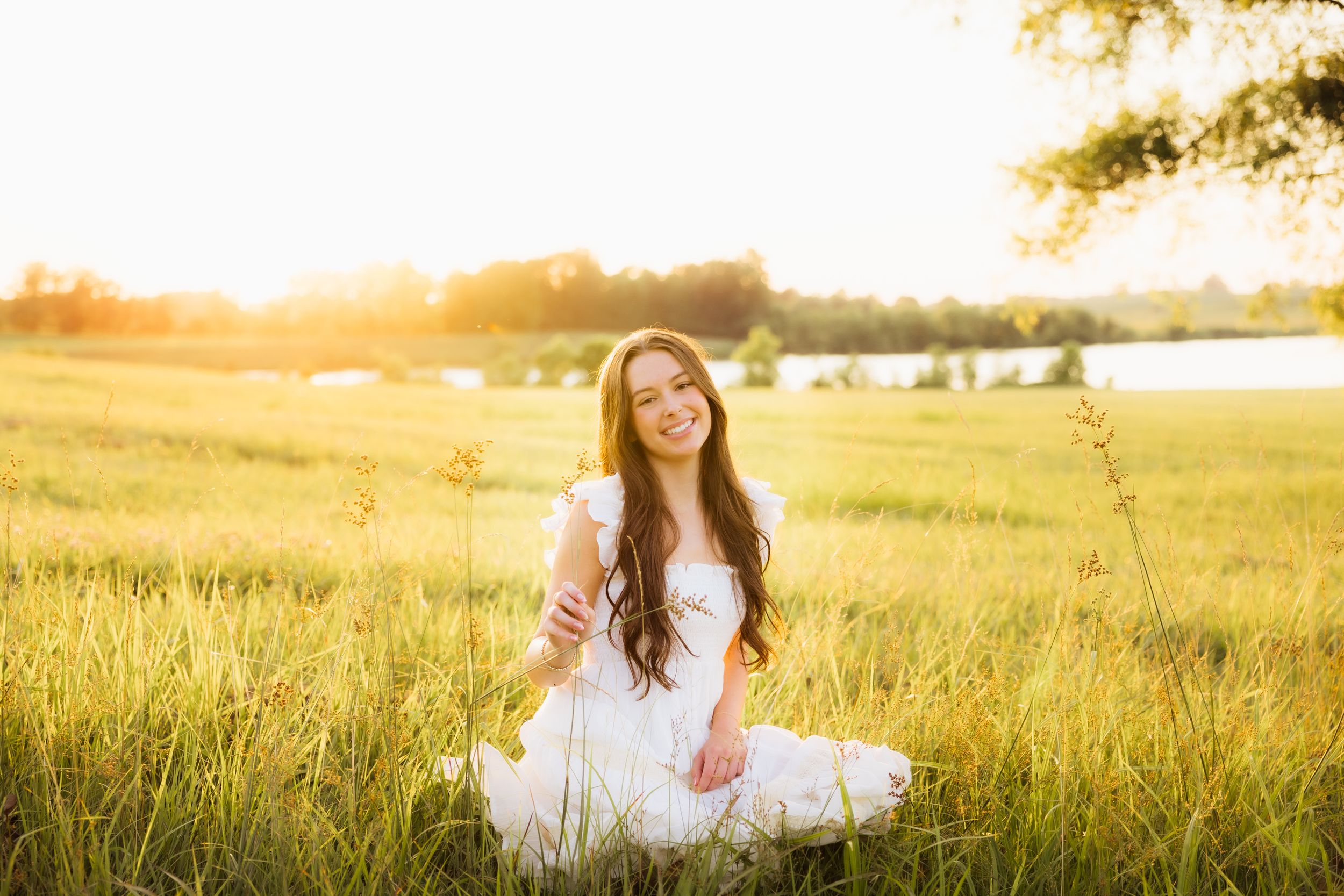 A senior in a white dress captured in a sunlit meadow during golden hour.
