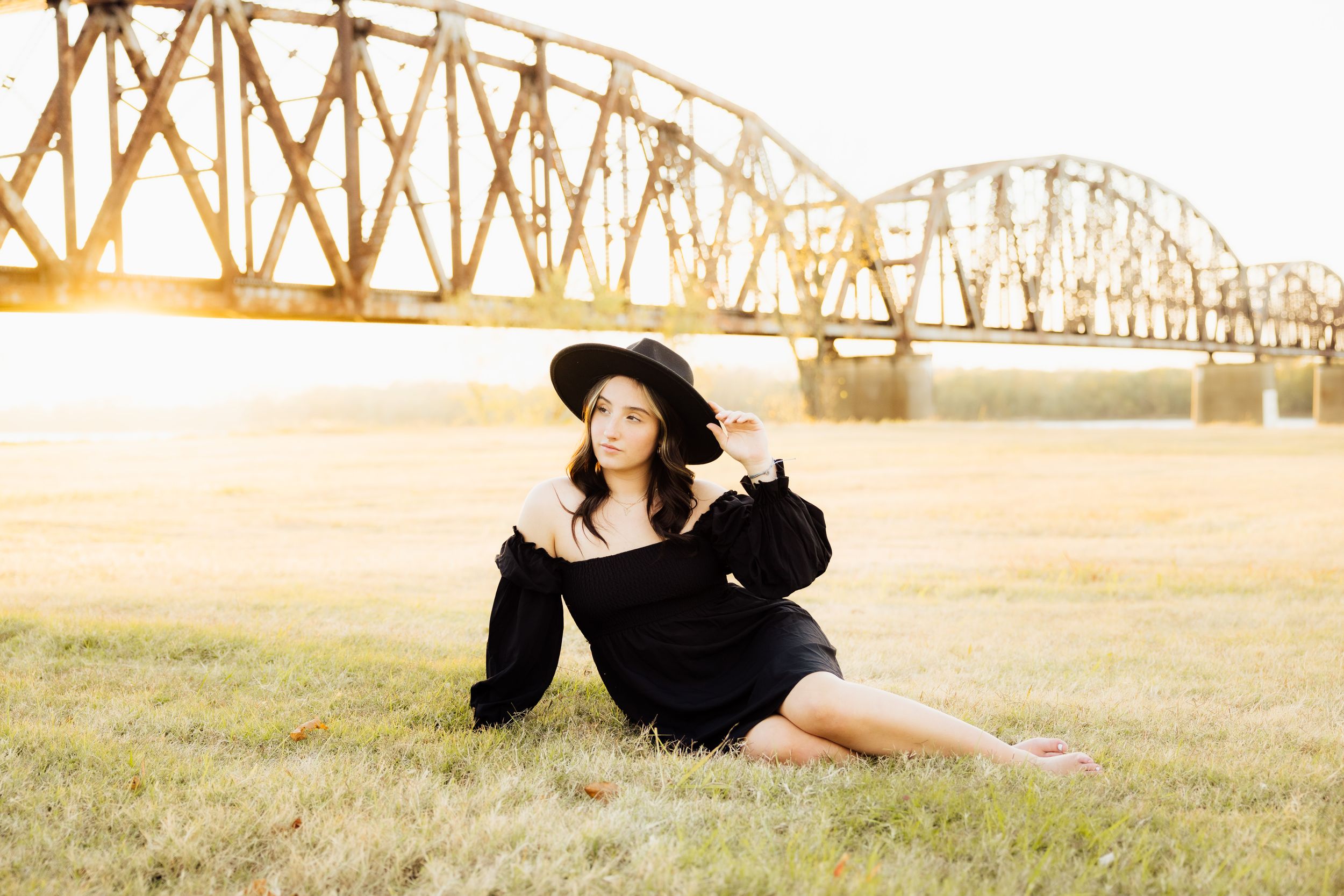 A senior in a black dress and wide brim hat ensemble photographed by a steel bridge at sunset.