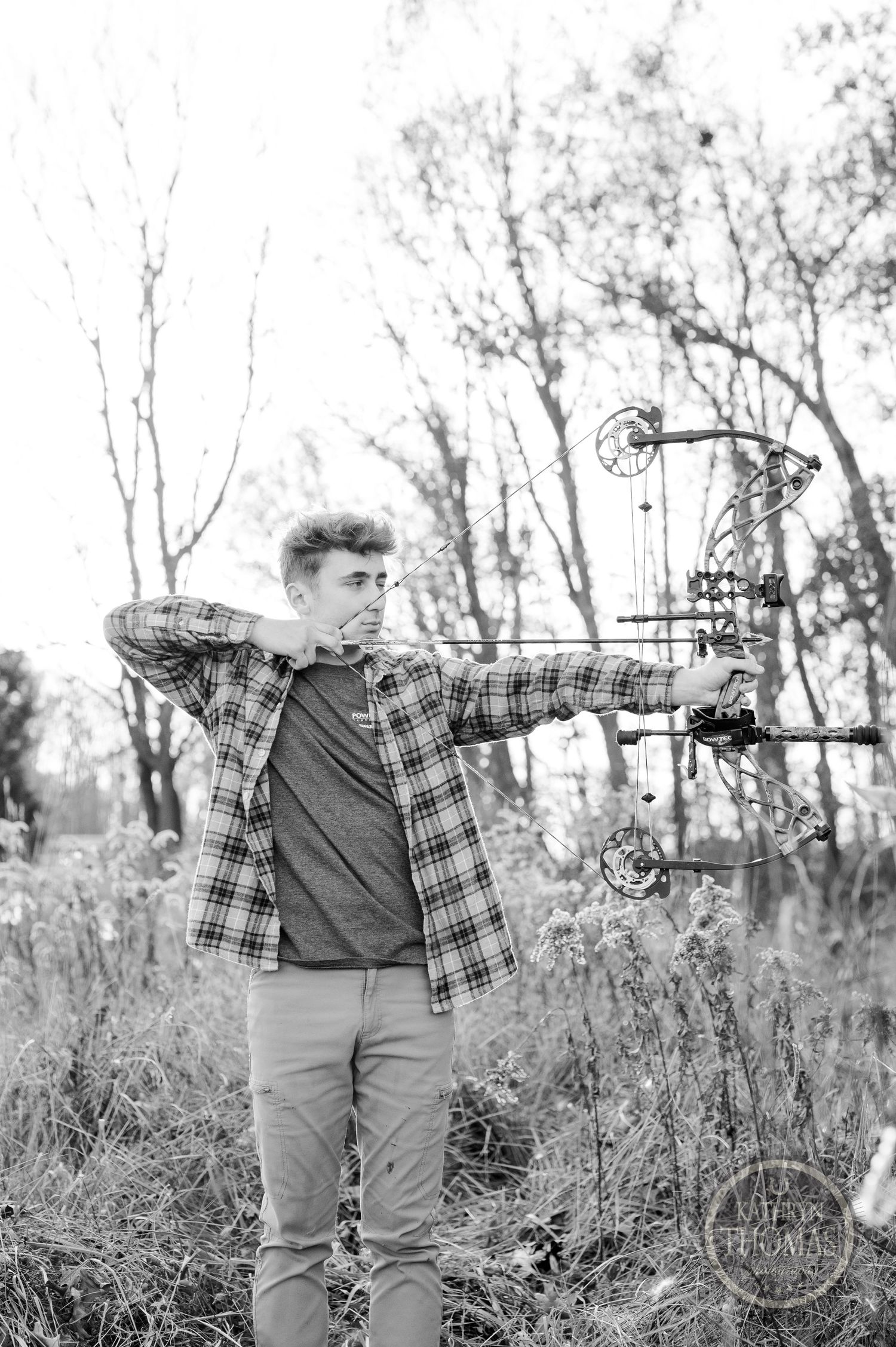 Black and white photo of archer drawing compound bow in wooded field.