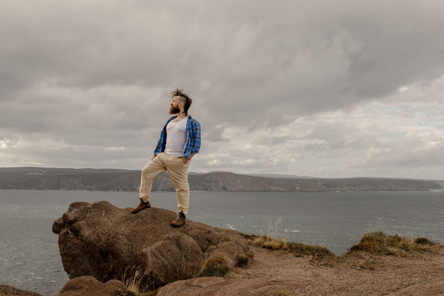 Figure in blue jacket and khaki pants stands on coastal rock formation under moody cloudy sky by ocean.