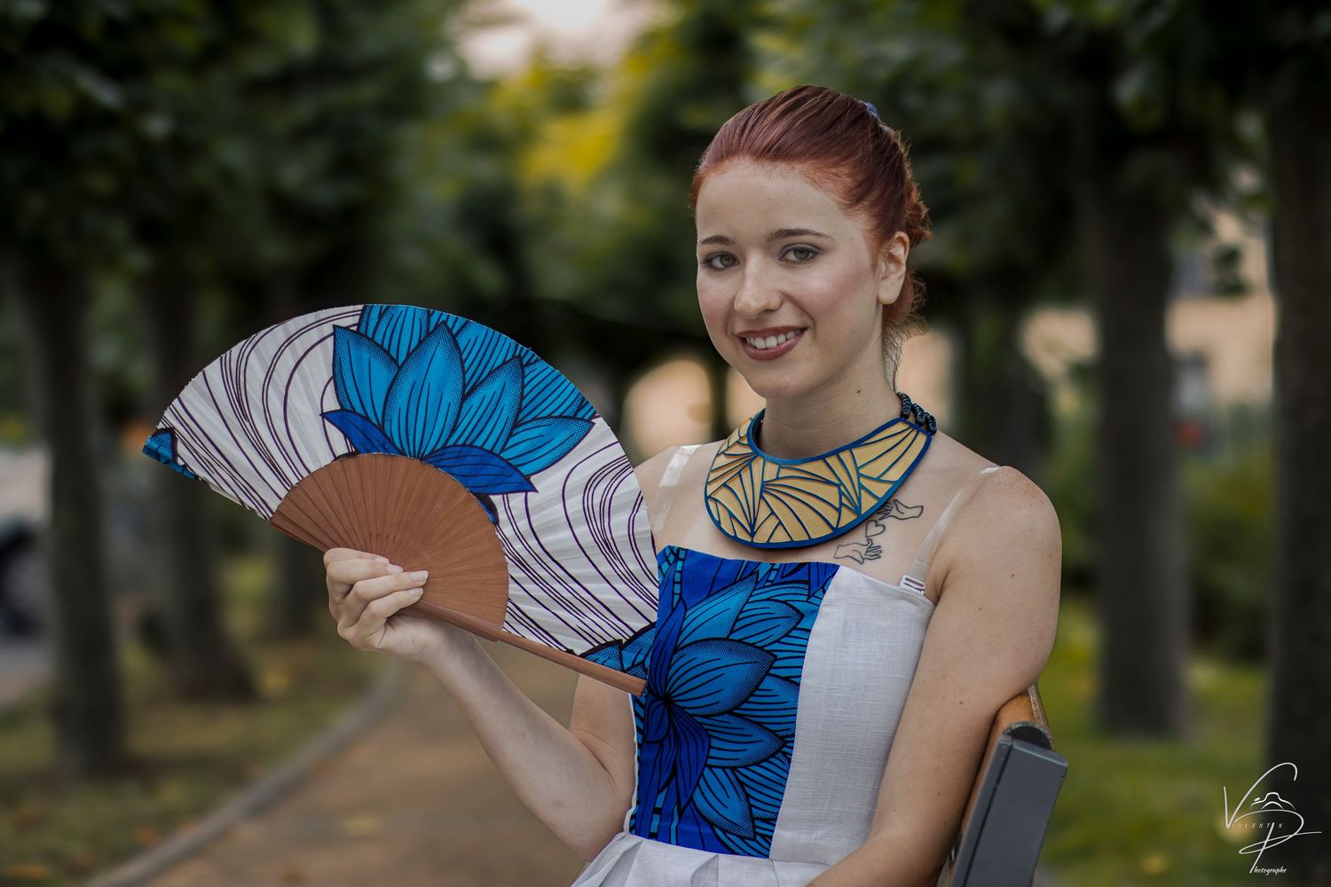 Person in costume poses with decorative blue and white fan in park setting.