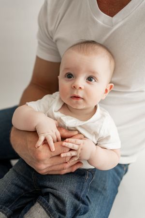 A series of sweet candid moments showing a baby being held on someone's lap in white clothing against a light background.