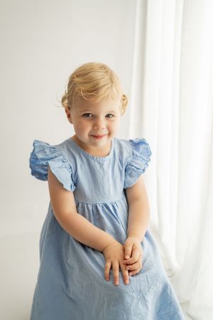 A joyful toddler wearing a light blue ruffled sleeve dress poses against a bright white background.