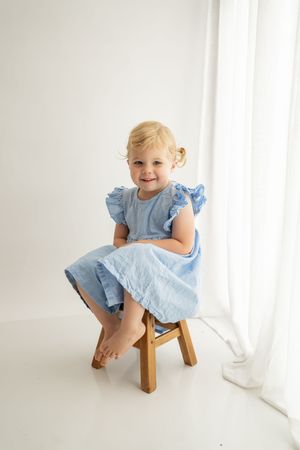 A small child in a pale blue dress sits playfully on a wooden stool near white curtains.