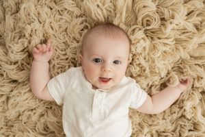 Little one lies on soft cream colored carpet while wearing white outfit and smiling at camera.