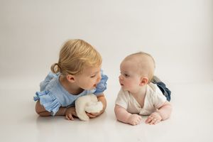 Two young children laying on their tummies on a white backdrop, wearing blue and white outfits.