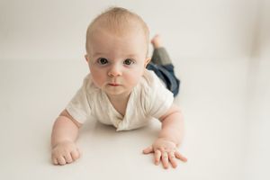 Small child in white outfit crawls and smiles on light colored background.