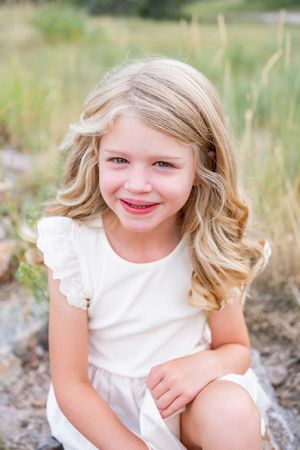 A young child in a white dress smiles while sitting outdoors among tall grass on a sunny summer day.
