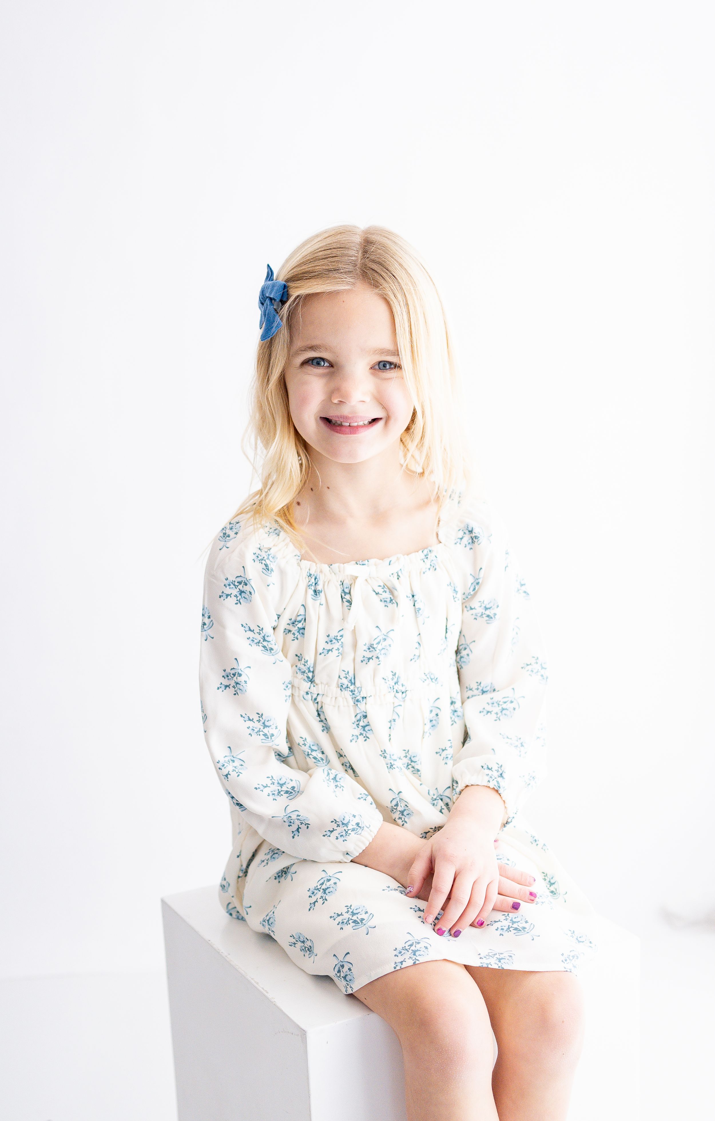 A young child wearing a white and blue floral dress sits on a white backdrop in three similar poses.